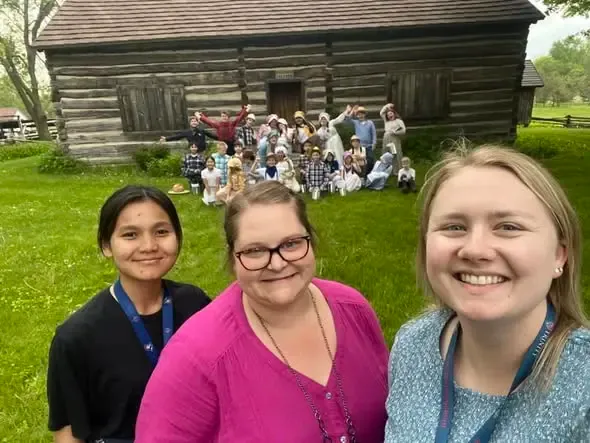 Three smiling chaperones with a class of schoolchildren in pioneer dress gathered in front of the log cabin