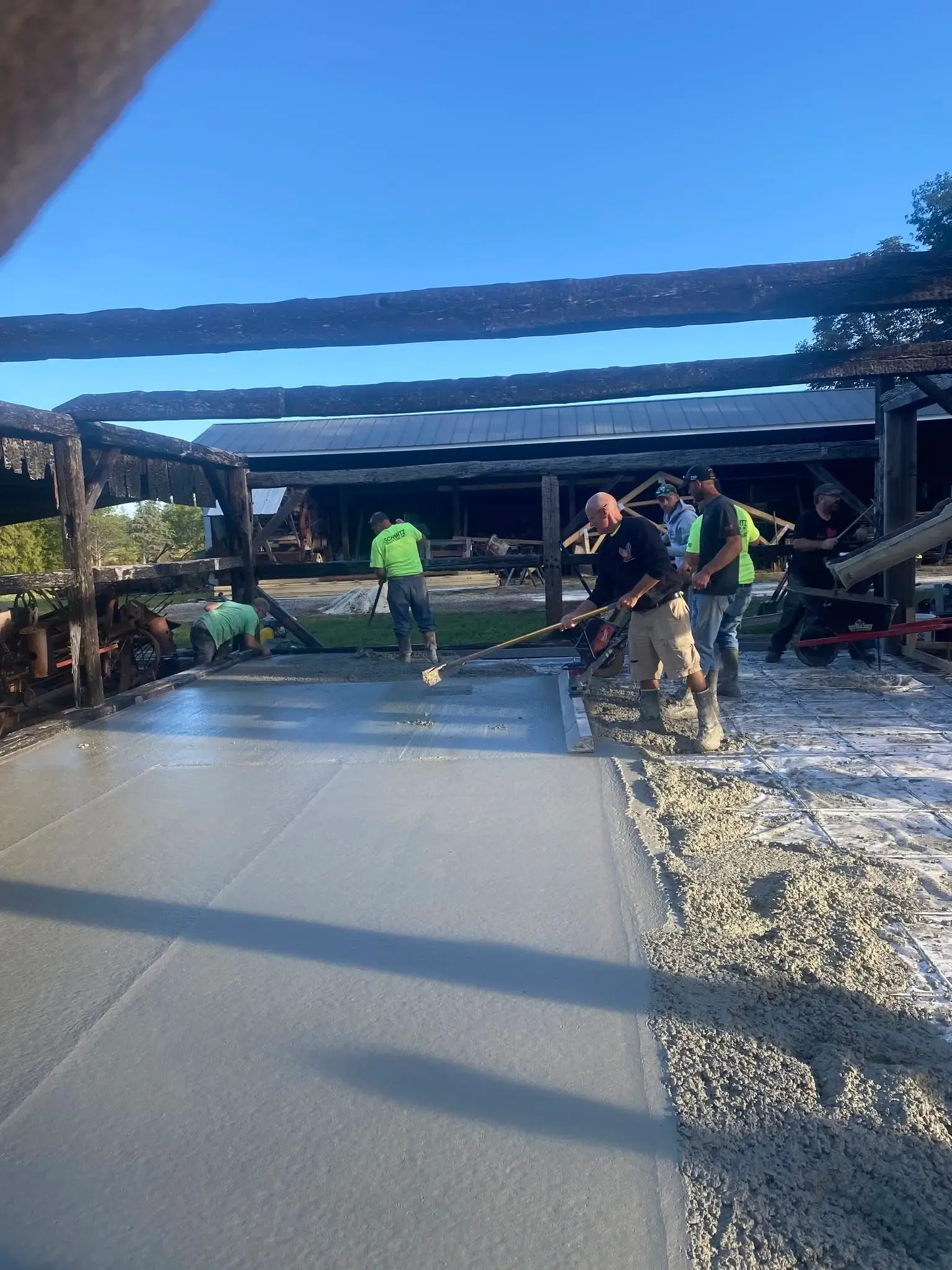 A worker guiding a power screed across the wet concrete while others rake it level beneath the shed's charred beams