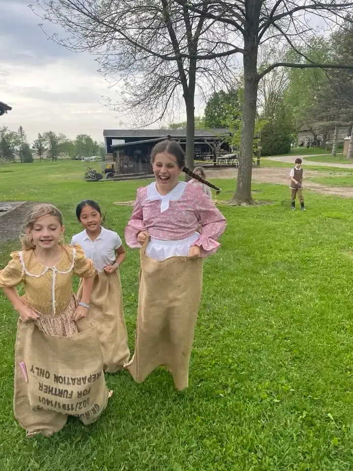 Girls in pioneer dresses laughing mid-hop through a burlap-sack race on the lawn