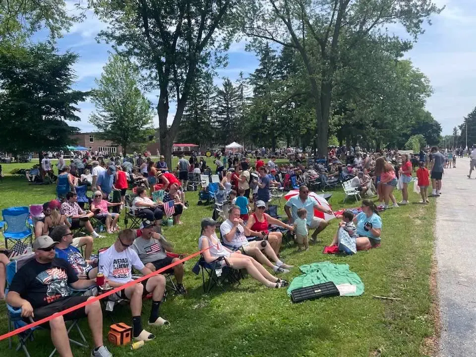 A crowd in lawn chairs lining the grassy parade route under big trees, waiting for the Freistadt 4th of July parade