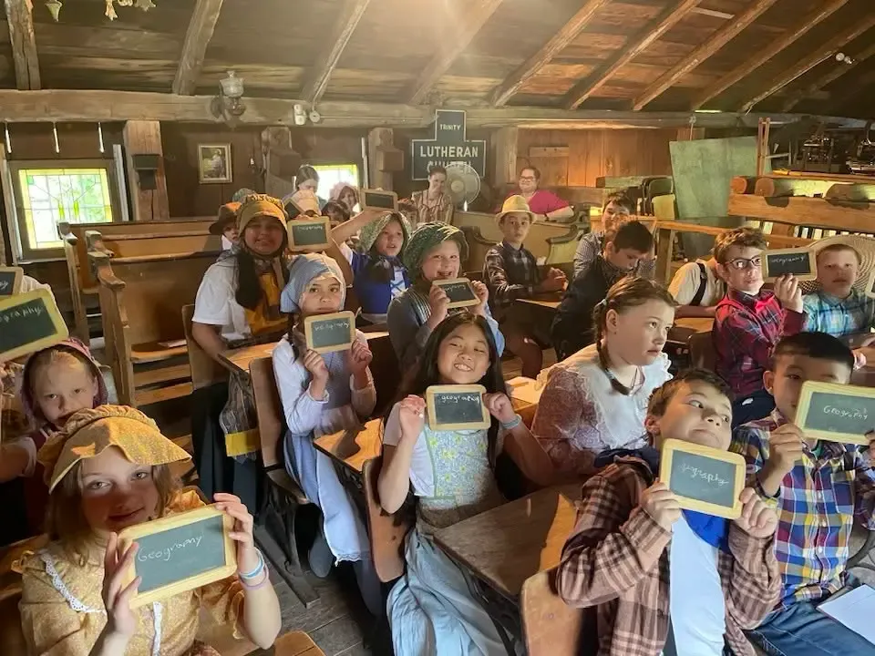 Schoolchildren in pioneer bonnets and overalls at wooden desks in the historic schoolhouse, holding up slates with “Geography” written in chalk