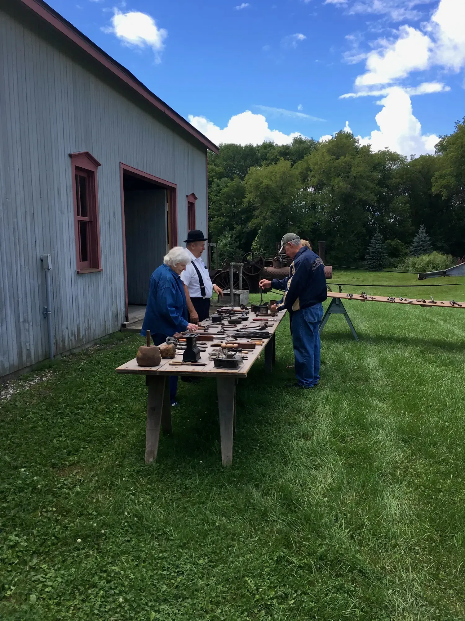Visitors browsing tables of antique hand tools outside the gray shed, the steam engine beyond