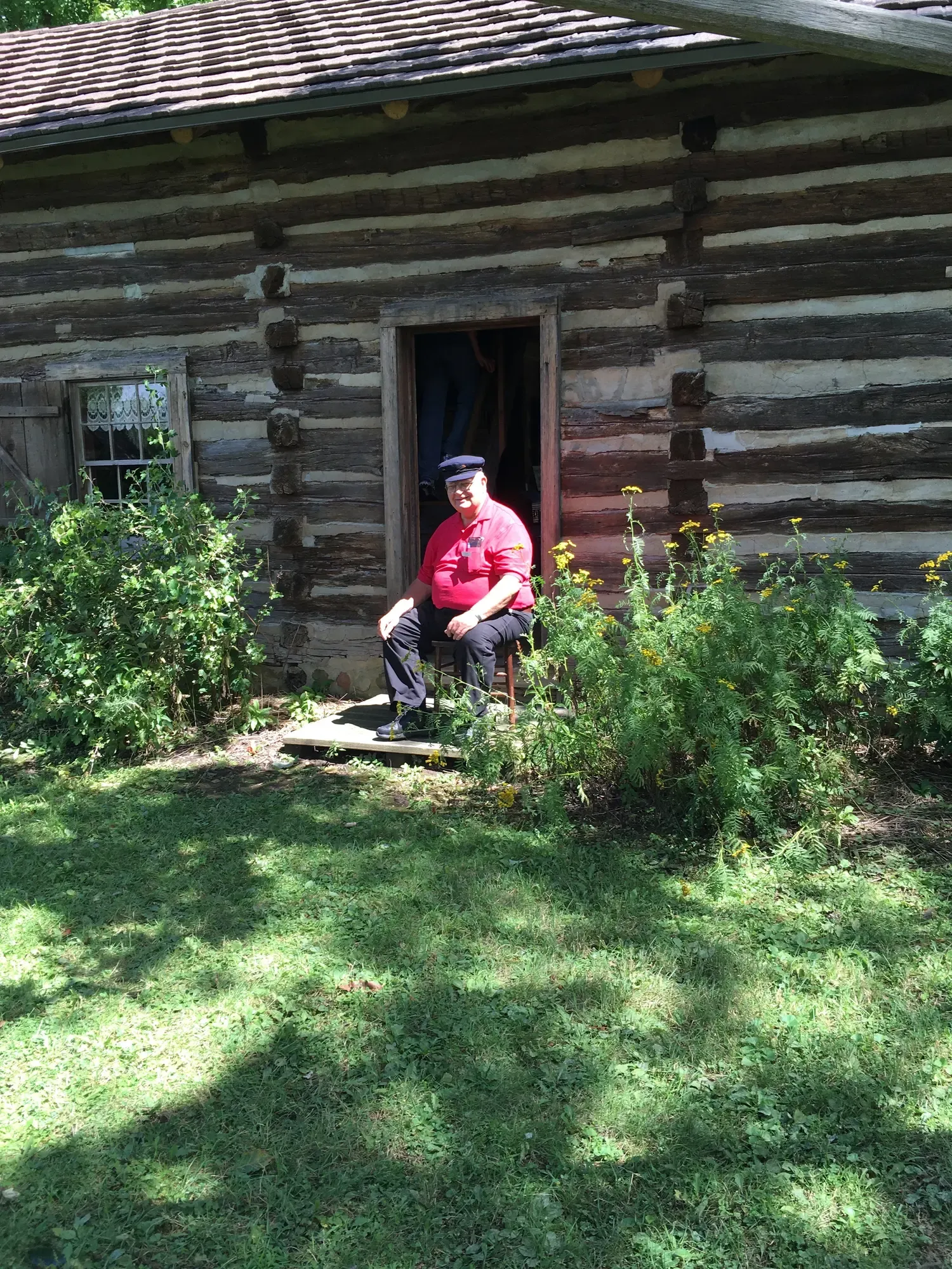 A volunteer in a period cap seated at the log cabin doorway among late-summer wildflowers