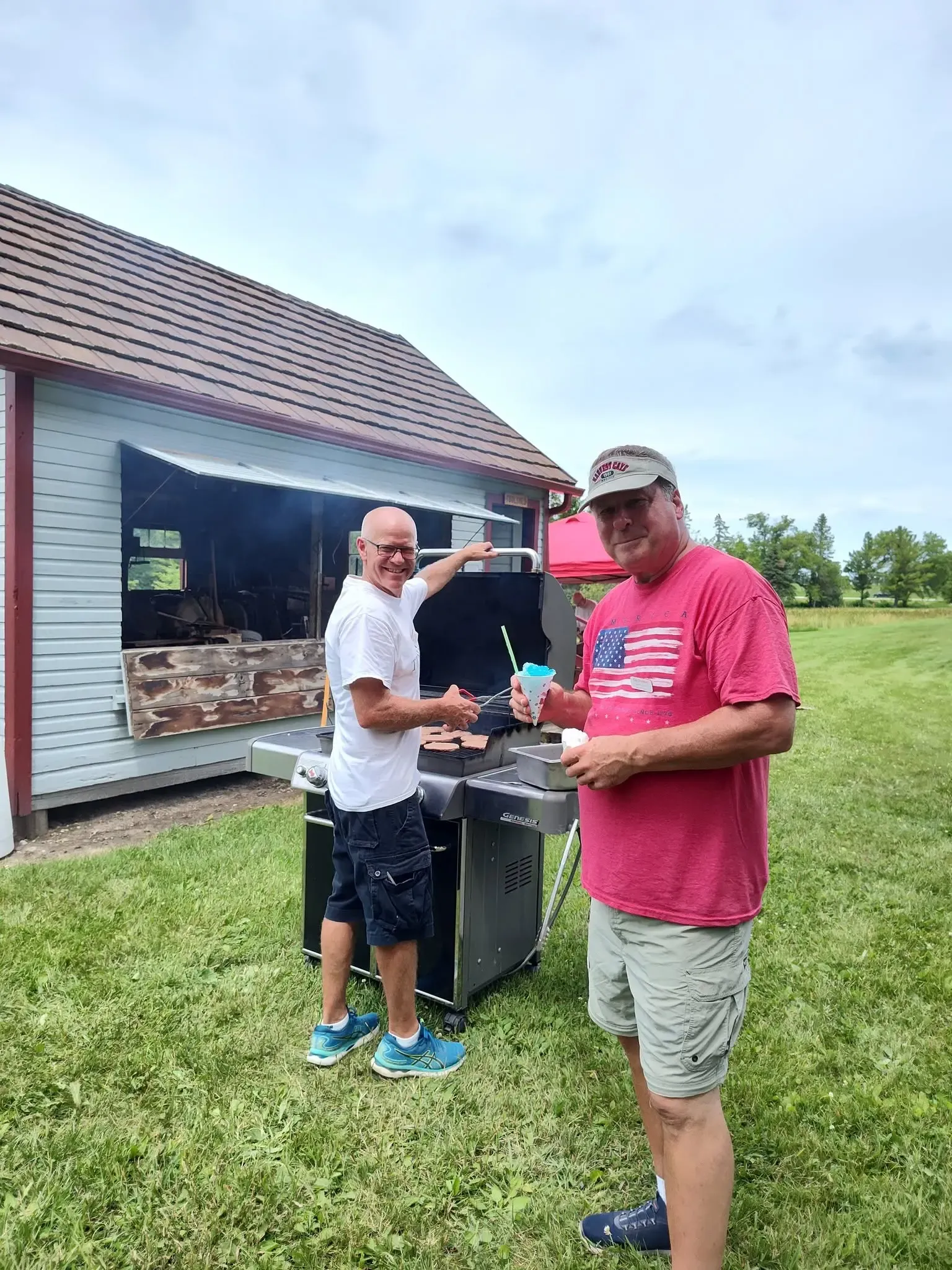 Two volunteers at the grill outside the tool shed, one with a blue snow cone in hand