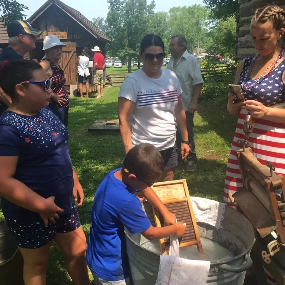 A boy scrubbing laundry on a washboard in a galvanized tub as Fourth of July visitors look on