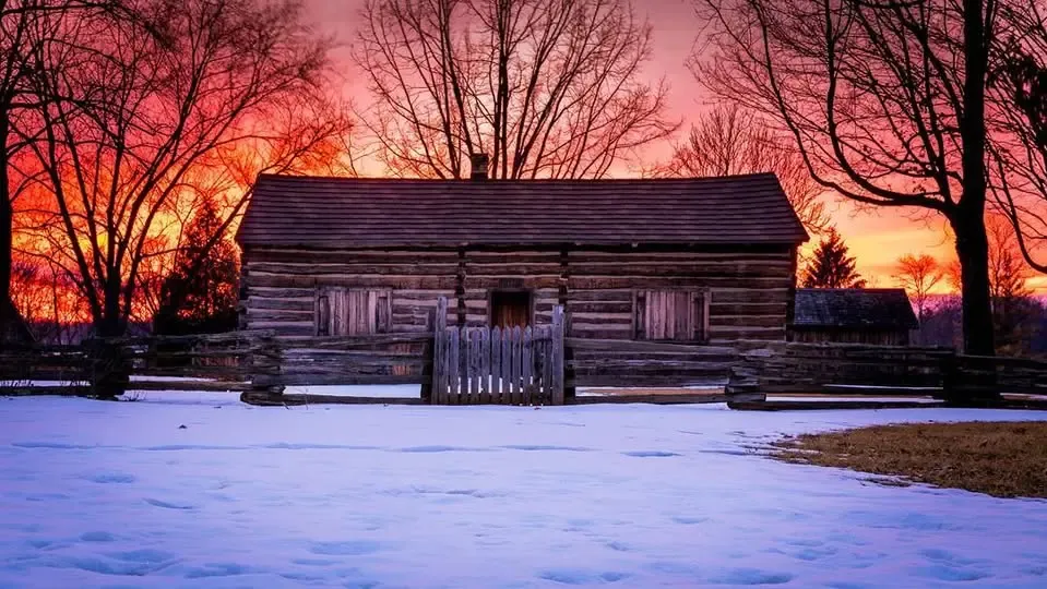 The log cabin at the historical site under snow at sunset, the sky glowing red and orange behind bare trees