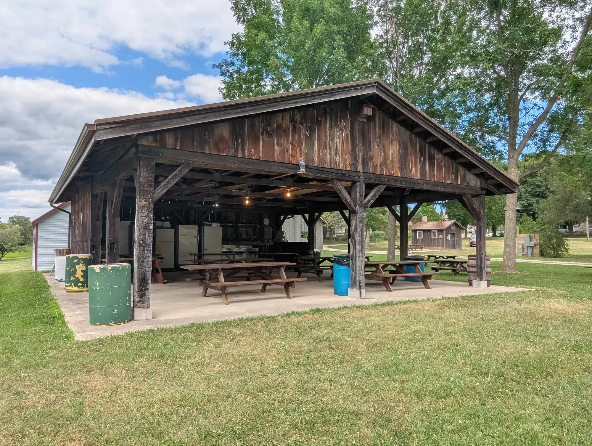 The timber-framed open picnic shelter with rows of picnic tables and kitchen equipment under its barnwood gable, big trees overhead