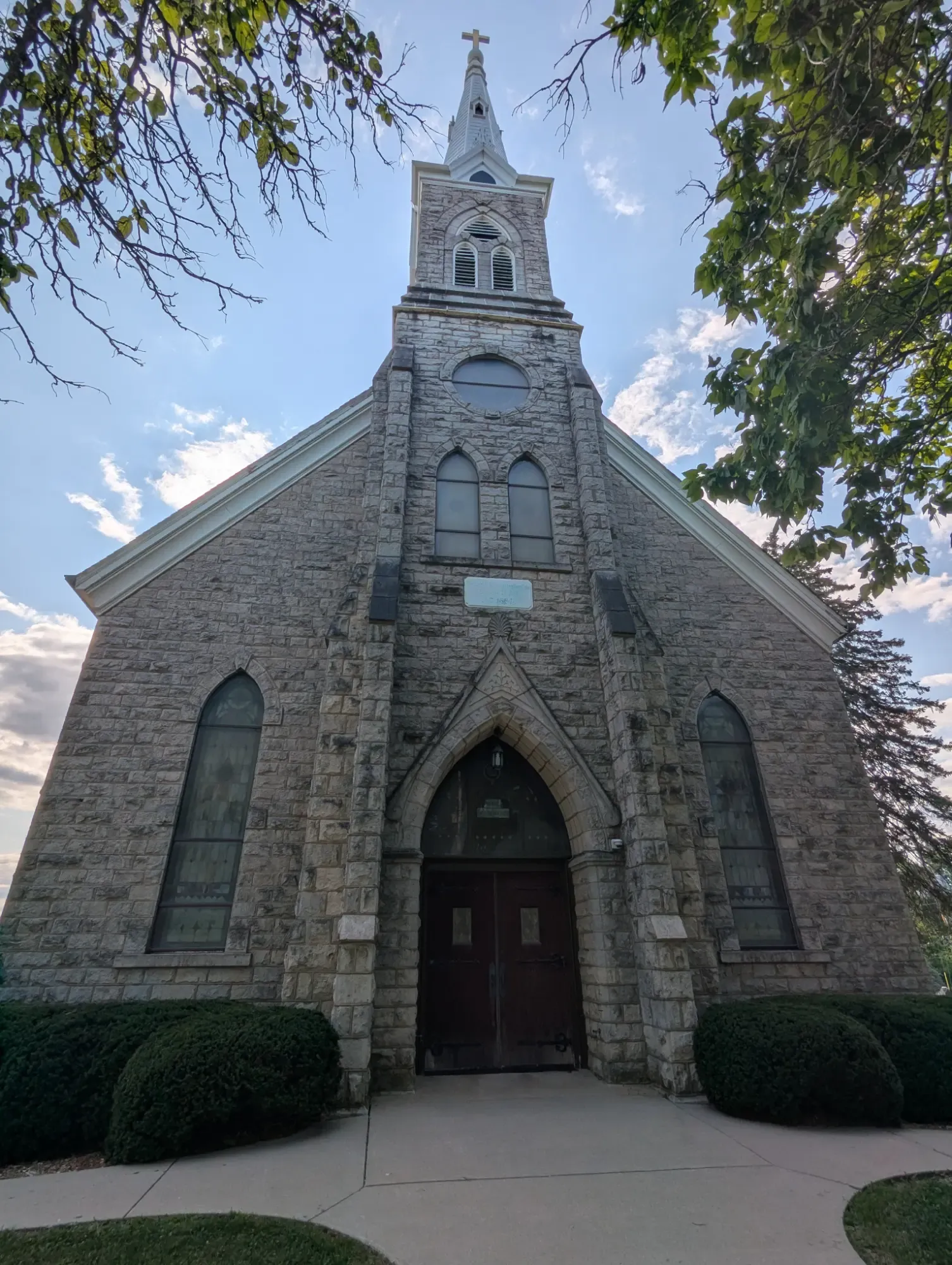 The stone front of the 1884 Trinity church, its steeple and cross rising between framing trees, arched stained-glass windows flanking the entrance
