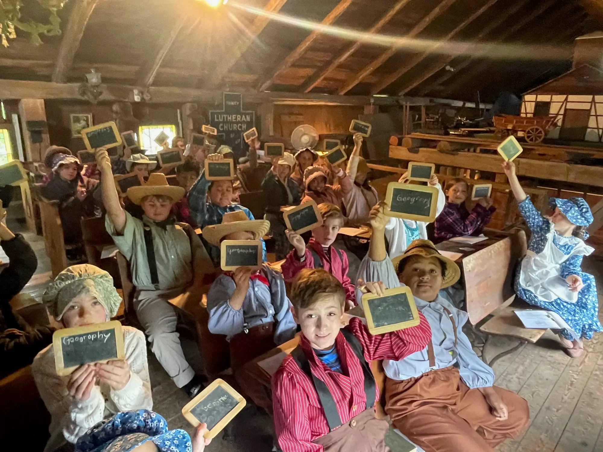 Schoolchildren holding up slates with 'Geography' chalked on them in the historic schoolroom