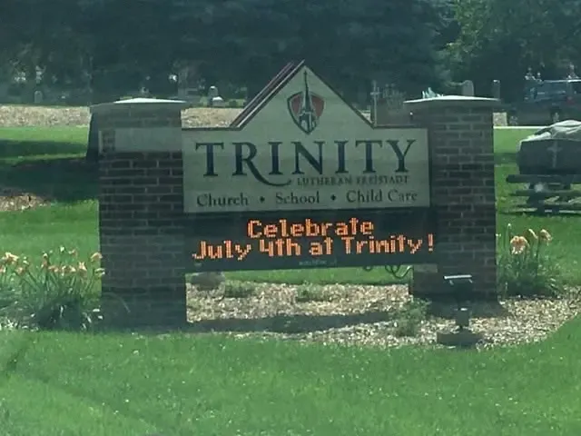 Brick Trinity sign on the church lawn reading “Celebrate July 4th at Trinity!” amid daylilies